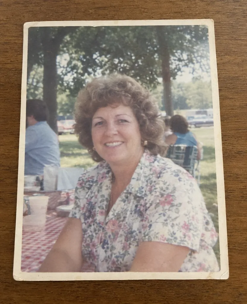 Faded 1980s print photo of a woman in a floral blouse smiling at a family picnic